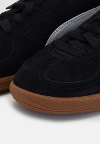 Gros plan sur des baskets en daim noir avec des semelles en caoutchouc marron texturé et des lacets noirs, montrant des coutures détaillées et la texture du tissu.