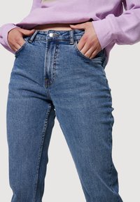 Jeans de mezclilla azul con cintura alta, corte recto, cinco bolsillos y costuras en contraste. Combinados con una blusa de manga larga de color púrpura claro.