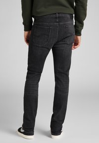 Man som bär svarta tajta jeans och svarta sneakers med vita sulor, står mot en ljusgrå bakgrund.