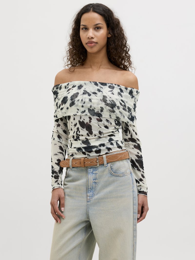 Top off-the-shoulder in tessuto leggero con macchie nere e marroni, abbinato a jeans di denim chiari a vita alta e una cintura marrone.