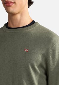 Grüner Strickpullover mit rundem Ausschnitt, strukturiertem Stoff und einer kleinen gestickten Norwegen-Flagge auf der Brust. Blaue Unterlage sichtbar.