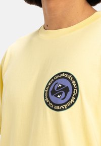 Camiseta de algodón amarilla con un logotipo gráfico redondo con acentos en azul y negro. Diseño de mangas cortas y cuello redondo. Textura suave.