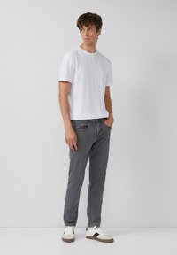 Jonge man staande in een wit T-shirt met korte mouwen, grijze jeans en witte sneakers met zwarte strepen tegen een effen grijze achtergrond.