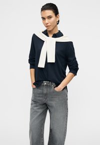 Πουκάμισο σε ναυτικό μπλε με κουμπιά και γυρισμένα μανίκια, συνδυασμένο με ψηλόμεσα γκρι τζιν. Μπεζ πουλόβερ δεμένο στον λαιμό προσθέτει ένα στοιχείο επιπέδων.