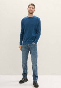 Suéter de punto azul con mangas largas, escote redondo, combinado con jeans azul claro y botas negras de tobillo, de pie sobre un fondo neutro.