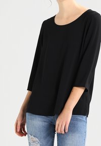 Svart långärmad tröja med rund halsringning, tillverkad av ett slätt tyg. Har en avslappnad passform och sidovents, matchad med ljusblå, slitna jeans.