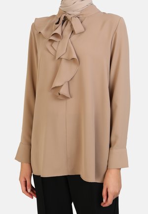 Beige blouse featuring a ruffled collar and tie detail, long sleeves, and a smooth texture, paired with black trousers.