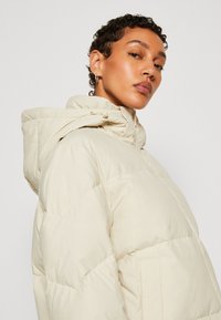 Beige péřová bunda s kapucí, s prošívaným designem, předním zipem a bočními kapsami. Lehce materiál, hladká textura.