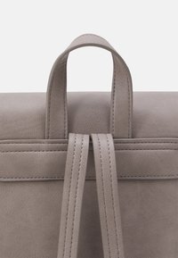 Primer plano de las correas y el asa de una mochila de cuero color topo, con costuras visibles en una superficie lisa y texturizada.