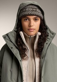 Olivovo zelená prešívaná bunda s kapucňou cez béžový štruktúrovaný sveter. Modelka má na hlave hnedú pletenú čiapku so vzorom. Detailný pohľad.
