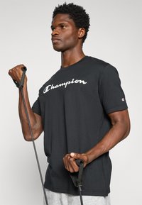Schwarzes Baumwoll-T-Shirt mit weißem "Champion"-Logo auf der Brust. Das Modell hält ein Widerstandsband. Graue Shorts sind unten sichtbar.