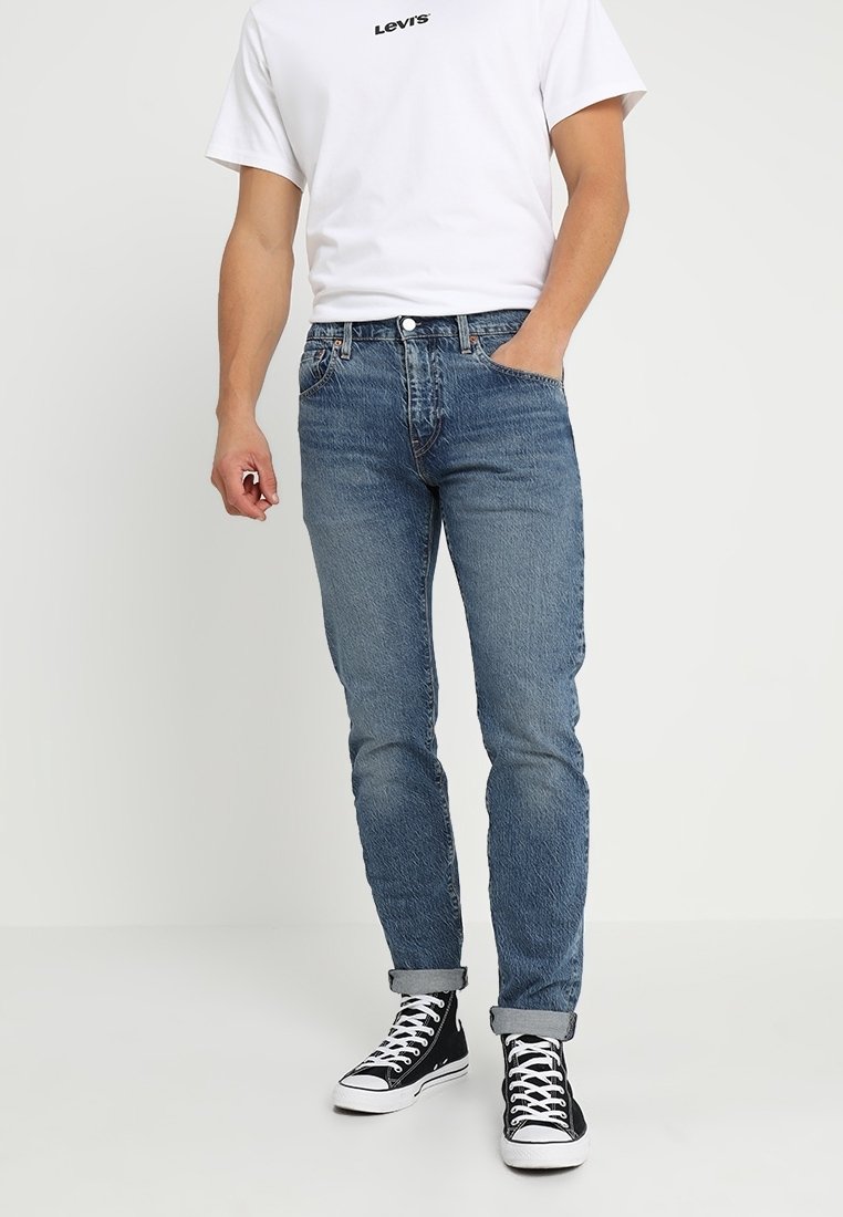 Blå denimjeans med smal passform, med subtil blekning och sydda detaljer, kombinerade med en enkel vit t-shirt och svarta höga sneakers.