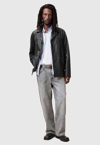 Mann mit langen Dreads, der eine schwarze Lederjacke, ein hellblaues Hemd, ein weißes T-Shirt, graue Jeans, schwarze Schuhe und einen braunen Gürtel trägt, aufrecht steht.