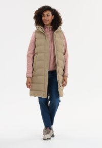 Beige puffer vest with a hood over a pink fleece top, paired with blue jeans and light-colored sneakers. Smooth, quilted texture.