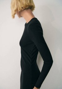 Black long-sleeve dress with a fitted silhouette, featuring smooth fabric and side ruching details, worn by a model in profile against a neutral background.