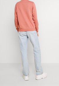 Pfirsichfarbener Pullover mit gerippten Bündchen und Saum, kombiniert mit hellblauen, geraden Jeans. Die Textur ist weich; die Sneakers sind weiß mit einer klobigen Sohle.