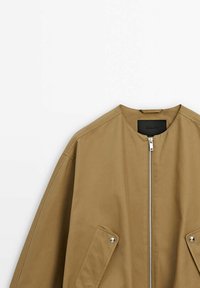 Veste bomber beige avec un col rond, une fermeture éclair à l'avant et deux poches latérales à pression. Texture de tissu lisse et léger.