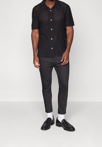 Chemise noire à manches courtes avec des motifs découpés complexes, associée à un pantalon ajusté sombre et des mocassins noirs. Chaussettes blanches visibles.