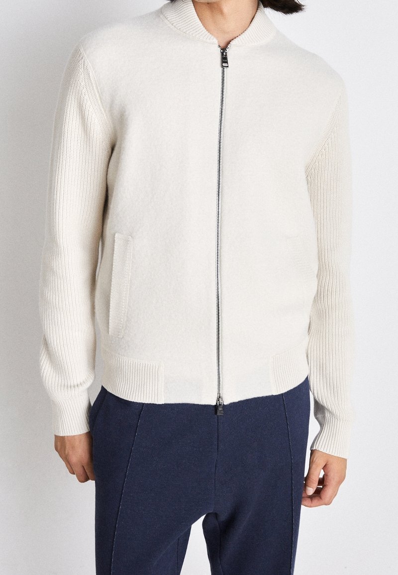 Personne portant un pull en maille blanc à fermeture éclair avec manches et col côtelés, associé à un pantalon bleu foncé, visible du cou aux hanches.