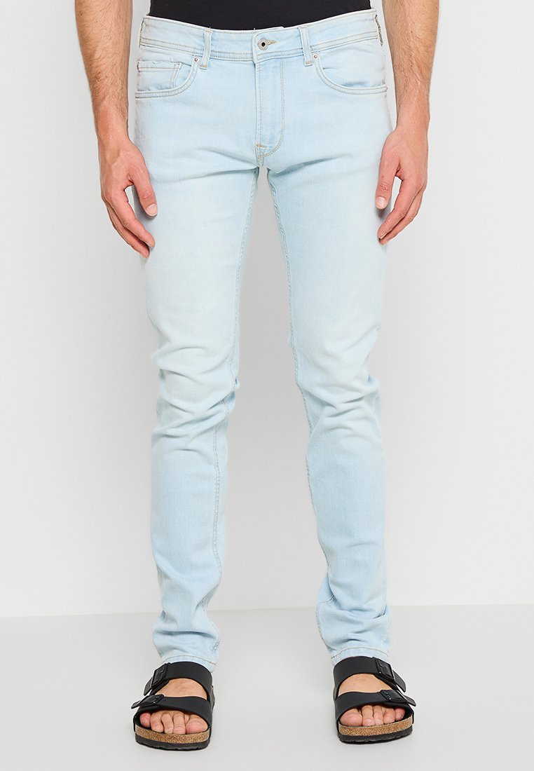 Helle blaue Skinny-Jeans aus Denim, mit vorderen Taschen und einem ausgewaschenen Look, kombiniert mit schwarzen Sandalen und einem schwarzen Oberteil.