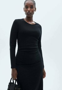 Black long-sleeve top with a rounded neckline, fitted design, and soft texture. Model holding a small black handbag.