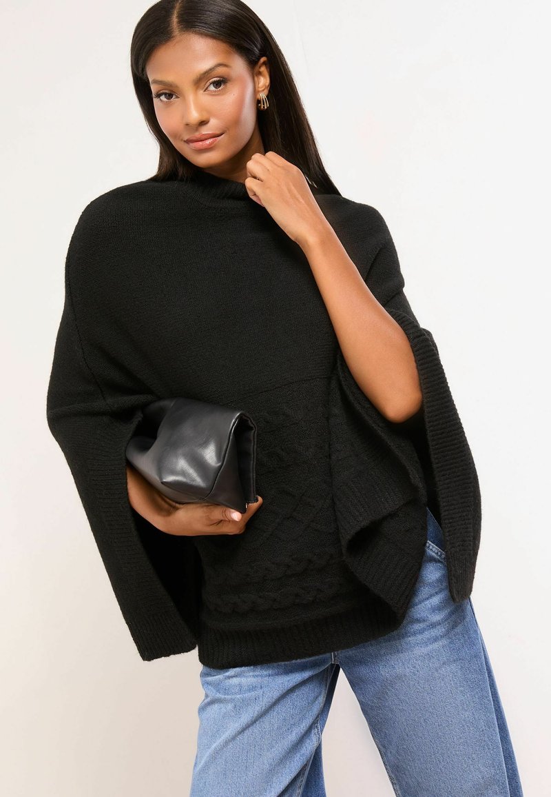 Poncho nero a maglia con collo alto, design testurizzato, maniche aperte e bordo a costine. Abbinato a una clutch nera e jeans blu.