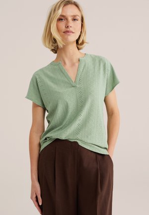 Mujer con blusa de manga corta texturizada color verde claro con escote en V y pantalones sueltos de cintura alta color marrón oscuro, de pie contra un fondo beige.
