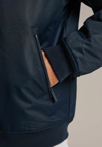 Granatowa kurtka bombka z zamkniętą kieszenią, ściągaczami przy mankietach i gładką teksturą materiału. Krój jest lekko zwężany w talii.