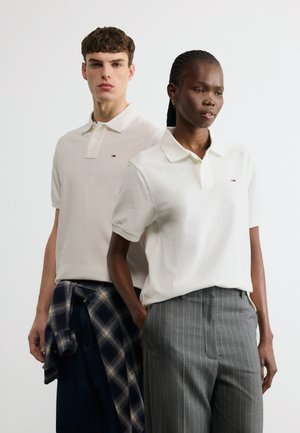 Δύο ενήλικες που φορούν λευκά πόλο μπλουζάκια, ο ένας με γκρι παντελόνι με ρίγες, ο άλλος με σκούρα τζιν και πουκάμισο καρό δεμένο στη μέση.