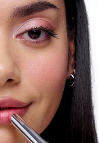 Gros plan sur un visage appliquant un rouge à lèvres rose dans un tube métallique argenté ; texture de peau lisse, rougeur naturelle et cheveux foncés visibles.