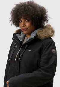 Black winter jacket with a fur-lined hood, textured grey interior, drawstring accents, and a logo patch on the upper arm.