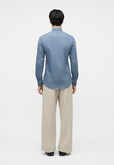 Άνδρας που στέκεται με την πλάτη προς το μέρος μας, φορώντας μπλε πουκάμισο με μακριά μανίκια και άνετο μπεζ παντελόνι, με μαύρα παπούτσια, πάνω σε ένα απλό φόντο.