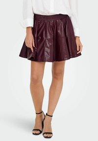 Mini-jupe en cuir bordeaux avec une taille élastique, un design évasé et une texture brillante, portée avec des escarpins noirs à lanières.