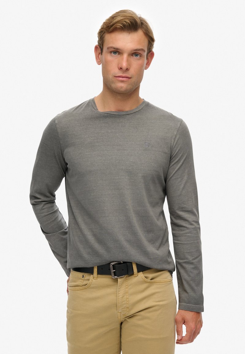 Langärmliges graues T-Shirt mit rundem Halsausschnitt und subtilen Logodetails. Kombiniert mit beigen Hosen und einem schwarzen Gürtel. Glatter Stofftextur.