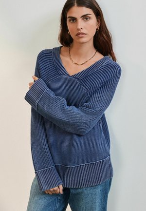 Blå stickad tröja med V-ringning, med olika randiga mönster, långa ärmar och ribbade muddar och nederkant; i kombination med jeans.