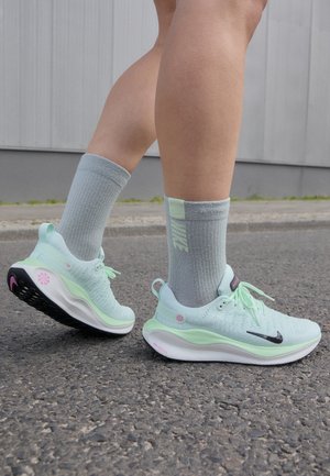 Nogi w jasnozielonych i białych butach do biegania z czarnymi logo Nike i szarych skarpetach Nike stojące na asfalcie przy szarej ścianie.