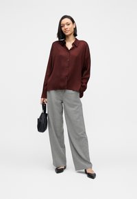 Chemise boutonnée à manches longues couleur bordeaux, associée à un pantalon large taille haute gris clair. Le mannequin tient un petit sac noir et porte des chaussures plates.