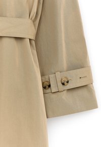 Manche de trench-coat beige présentant une texture lisse, un poignet ceinturé avec deux boutons ronds et un détail de coupe ajustée.