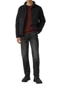 Czarna kurtka z pikowanym przodem, czerwona sweter, ciemne dżinsy i czarne buty. Kurtka ma zamek błyskawiczny i kieszeń na piersi.