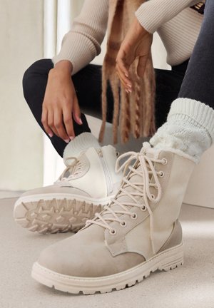 Beige Stiefeletten mit Wildlederfinish, Schnürdesign und strukturiertem Gummisohlen. Der linke Stiefel verfügt über ein seitliches Reißverschlussdetail.