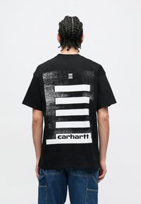 Camiseta negra de algodón con un gran diseño gráfico en la espalda, con franjas horizontales y el logo "Carhartt" en blanco.