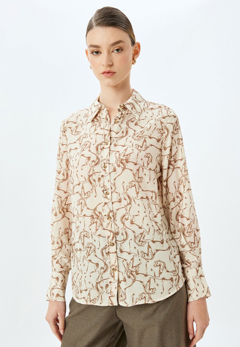 Patroonblouse in beige met bruine paardenmotieven, met een knoopsluiting aan de voorkant, kraag en lange mouwen van lichte stof.