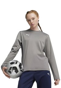 Maglietta sportiva grigia a maniche lunghe con scollatura rotonda, caratterizzata da un logo bianco. La persona tiene un pallone da calcio a motivi bianco e nero.