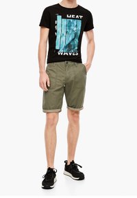 T-shirt noire avec un design graphique bleu affichant "HEAT WAVES". Short vert olive avec des ourlets retournés, associé à des baskets noires.