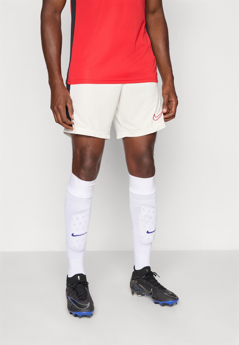 Nike sporta komplekts ar sarkanu krekli, gaišiem šortiem, baltām zeķēm ar zilu akcentu un melniem futbola apaviem ar zilu detalizāciju.