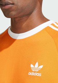 Camiseta naranja de Adidas con cuello blanco y tres rayas. Presenta un logotipo bordado en blanco en el pecho. Textura de algodón suave.
