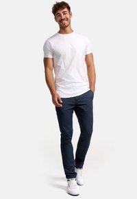 Weißes T-Shirt mit kurzen Ärmeln, kombiniert mit dunkeln, schmal geschnittenen Jeans. Das Model trägt weiße Sneaker mit bunten Akzenten. Glatte Stoffstruktur.