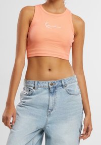 Žena v bezramínkovém broskvovém topu s bílým nápisem a v světle modrých džínech s vysokým pasem na jednobarevném pozadí.