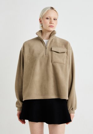 Beiger Fleecepullover mit hohem Kragen, Halbreißverschluss und einer Tasche auf der Vorderseite. Weiche Textur, oversized Schnitt und voluminöse Ärmel. Darunter ein schwarzer Rock.