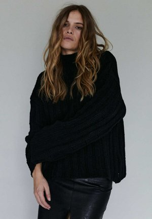 Czarny dziergany sweter z wysokim kołnierzem, o prążkowanej fakturze i oversizowych rękawach. Połączony z czarną skórzaną spódnicą.